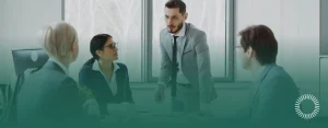 A man in a gray suit leans over a conference table speaking to three colleagues during a tense discussion, Optima Juris logo bottom right.